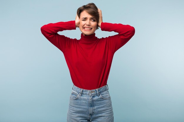 Giovane signora stanca infastidita in maglione rosso e jeans che chiude tristemente le orecchie con le mani su sfondo blu isolato