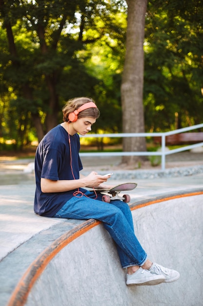 Giovane skater in cuffie arancioni mentre usa premurosamente il cellulare con lo skateboard vicino al moderno skatepark