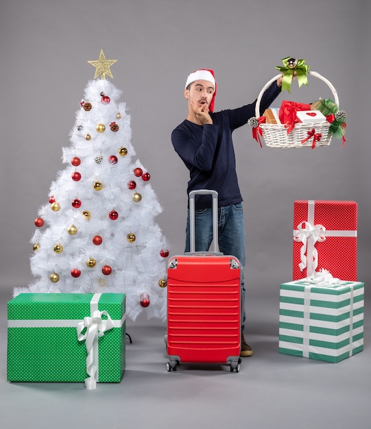 giovane sorpreso con cappello santa guardando cesto regalo vicino albero di Natale su grigio