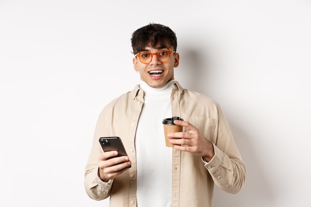 Giovane sorridente allegro con gli occhiali che beve caffè e tiene in mano uno smartphone, che guarda eccitato alla telecamera, in piedi su sfondo bianco.