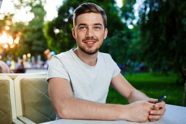 Giovane sorridente bello che si siede al tavolo nel distogliere lo sguardo all'aperto del telefono della tenuta del caffè.