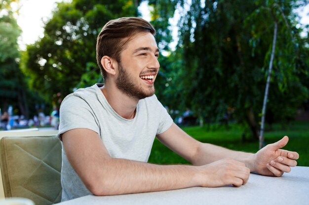 Giovane sorridente bello che si siede alla tavola in caffè all'aperto e distogliere lo sguardo.