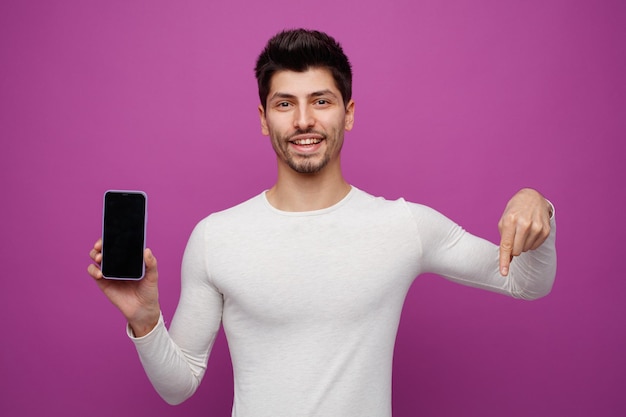 Giovane sorridente che guarda la fotocamera che mostra il telefono cellulare alla fotocamera rivolto verso il basso isolato su sfondo viola