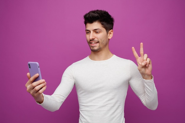 Giovane sorridente che tiene il telefono cellulare che mostra il segno di pace che prende selfie isolato su fondo porpora
