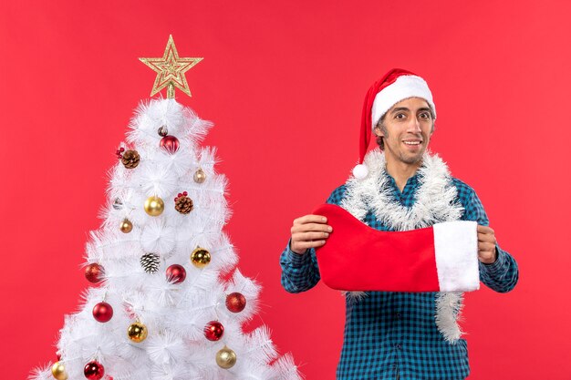 Giovane sorridente energico con il cappello di Babbo Natale in una camicia spogliata blu e che tiene il calzino di Natale