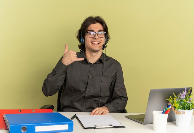 Giovane sorridente ufficio lavoratore uomo sulle cuffie in vetri ottici si siede alla scrivania con strumenti di office utilizzando laptop gesti telefono segno della mano