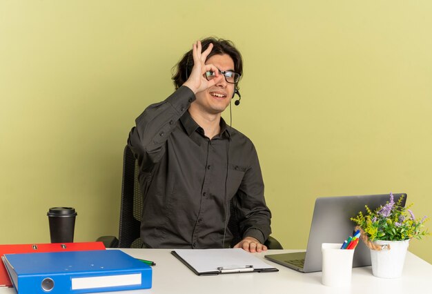 Giovane sorridente ufficio lavoratore uomo sulle cuffie in vetri ottici si siede alla scrivania con strumenti per ufficio utilizzando laptop guarda attraverso le dita isolate su sfondo verde con spazio di copia