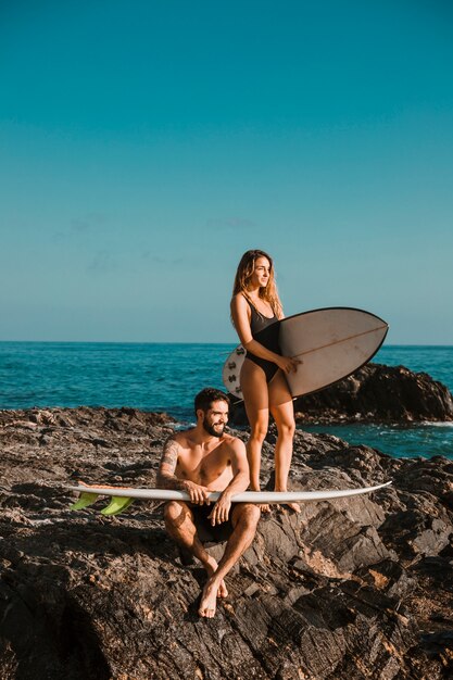 Giovane sorridente uomo e donna con tavole da surf sulla roccia vicino al mare