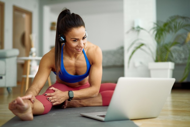 Giovane sportiva che si allunga sul pavimento mentre si utilizza il laptop e seguendo le istruzioni su Internet