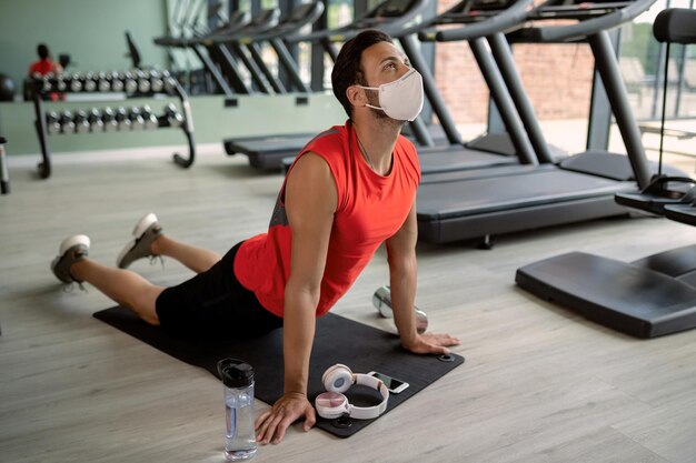 Giovane sportivo che fa esercizio di rilassamento mentre indossa una maschera protettiva in palestra a causa dell'epidemia di coronavirus