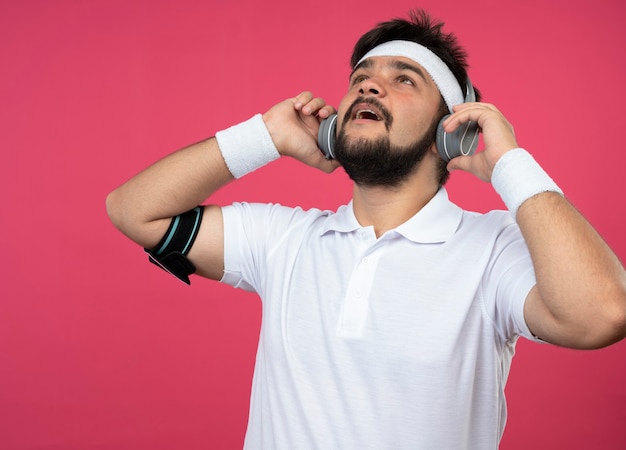 Giovane sportivo sorpreso che osserva in su che indossa la fascia e il braccialetto con le cuffie e la fascia del braccio del telefono