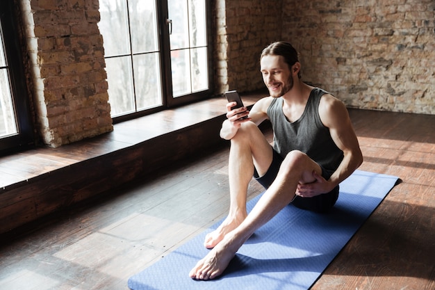 Giovane sportivo sorridente che per mezzo del telefono cellulare mentre sedendosi