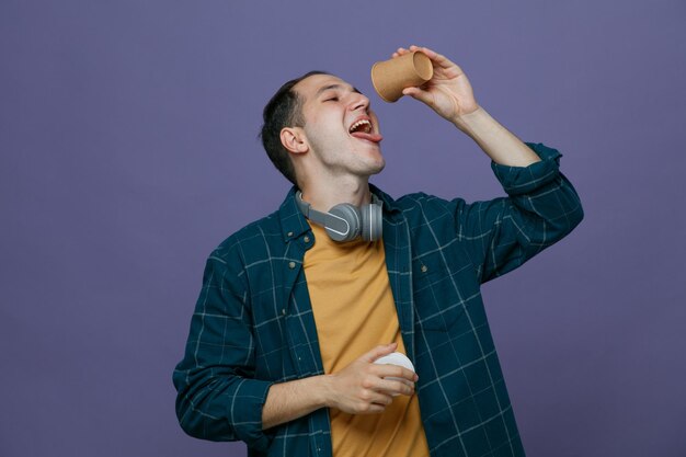 giovane studente assetato di sesso maschile che indossa le cuffie intorno al collo tenendo una tazza di caffè di carta sopra la bocca e il suo cappuccio in un'altra mano guardando nella tazza con la bocca aperta che mostra la lingua isolata su sfondo viola