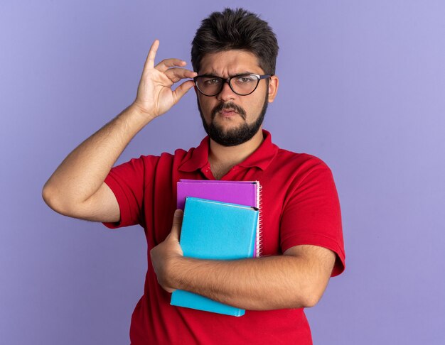 Giovane studente barbuto con una polo rossa che tiene in mano libri con gli occhiali da vicino con una faccia seria in piedi sul muro blu blue