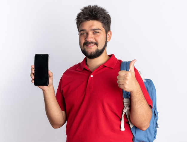 Giovane studente barbuto in polo rossa con zaino che mostra smartphone sorridente fiducioso che mostra i pollici in su in piedi sul muro bianco