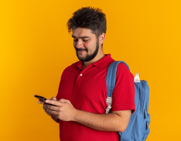 Giovane studente barbuto in polo rossa con zaino che tiene in mano lo smartphone e lo guarda sorridendo allegramente felice e positivo in piedi sul muro arancione