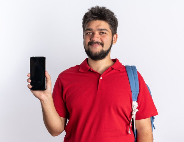 Giovane studente barbuto in polo rossa con zaino che tiene in mano uno smartphone che guarda in piedi