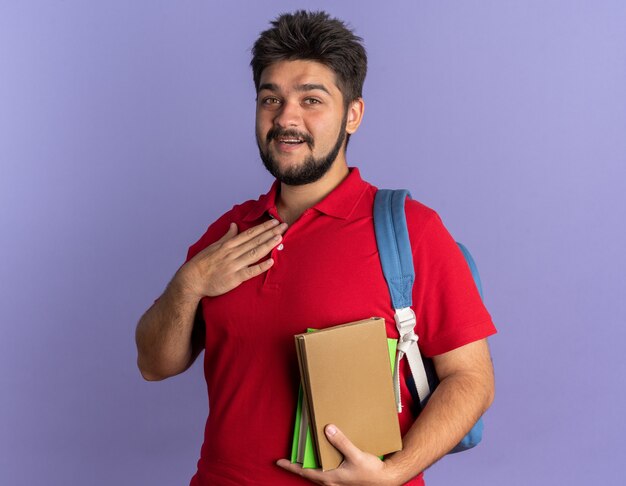 Giovane studente barbuto in polo rossa con zaino che tiene libri tenendo la mano sul petto sentendosi grato sorridendo allegramente in piedi sul muro blu