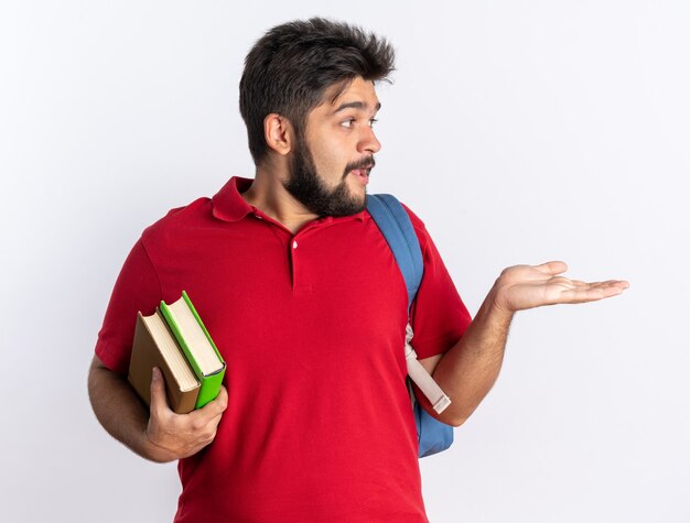 Giovane studente barbuto in polo rossa con zaino che tiene quaderni che guarda da parte sorpreso presentando con il braccio della mano copia spazio in piedi sul muro bianco