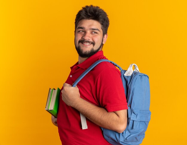 Giovane studente barbuto in polo rossa con zaino che tiene quaderni che guarda da parte sorridente fiducioso felice e positivo in piedi sul muro arancione orange