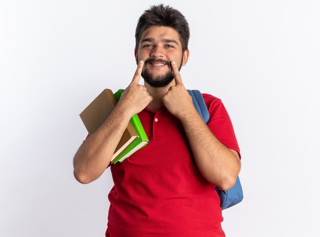 Giovane studente barbuto in polo rossa con zaino che tiene quaderni che puntano con le dita al suo grande sorriso in piedi sul muro bianco