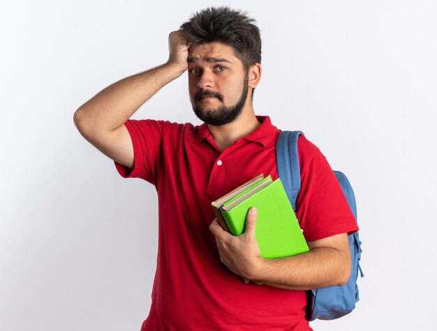 Giovane studente barbuto in polo rossa con zaino che tiene quaderni confusi con la mano sulla testa per errore in piedi sul muro bianco