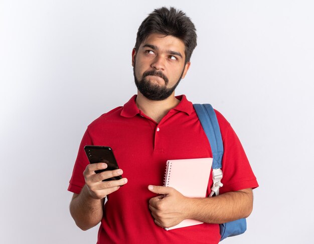 Giovane studente barbuto in polo rossa con zaino che tiene smartphone e taccuino alzando lo sguardo perplesso in piedi sul muro bianco