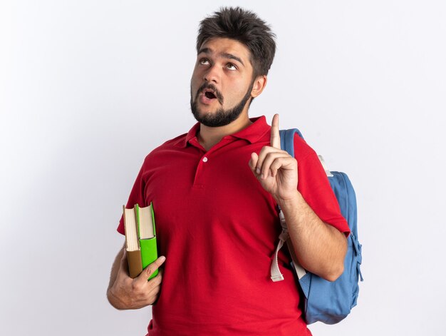 Giovane studente barbuto in polo rossa con zaino con in mano i taccuini alzando lo sguardo sorpreso mostrando il dito indice con una nuova idea in piedi