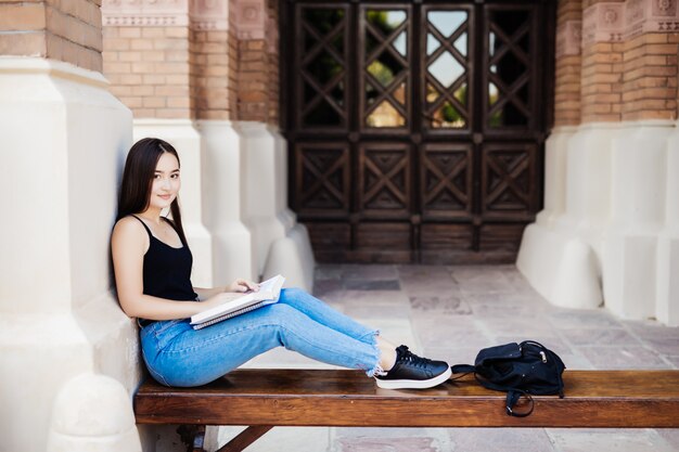 Giovane studente di college asiatico che legge un libro che si siede sul banco al campus universitario