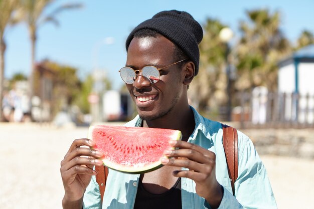 Giovane studente maschio afroamericano allegro d'avanguardia d'avanguardia che indossa le tonalità di lente a specchio alla moda e cappello nero che mangiano anguria matura mentre trascorrendo giorno di estate con gli amici fuori sulla spiaggia della città