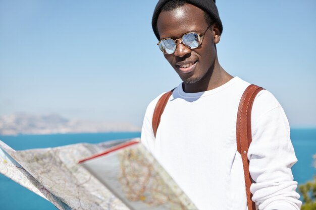 Giovane studente maschio afroamericano allegro in occhiali da sole con lenti a specchio alla ricerca di nuovi luoghi e punti di riferimento da visitare sulla mappa cartacea nelle sue mani durante un viaggio all'estero durante le vacanze estive