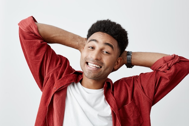 Giovane studente maschio amercan abbronzato allegro felice con l'acconciatura afro in maglietta bianca che sorride con i denti, tenentesi per mano dietro la testa che posa per la foto di università di grduation.