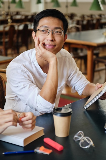 Giovane studente maschio asiatico attraente in occhiali che studia sognante nella biblioteca dell'università