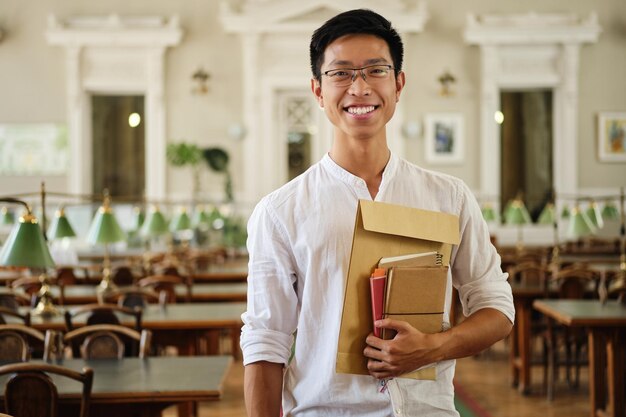 Giovane studente maschio asiatico sorridente in occhiali con taccuini guardando con gioia a porte chiuse nella biblioteca dell'università