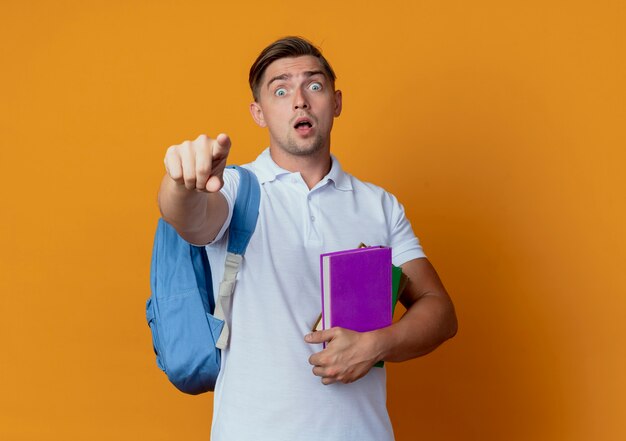 Giovane studente maschio bello sorpreso che porta il libro della tenuta della borsa posteriore e punti alla macchina fotografica isolata sull'arancia