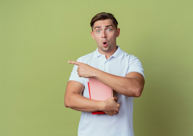 Giovane studente maschio bello sorpreso che tiene libri e punti a lato isolato sulla parete verde oliva con lo spazio della copia