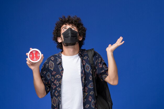 Giovane studente maschio di vista frontale che porta maschera nera con gli orologi della holding dello zaino sui precedenti blu.