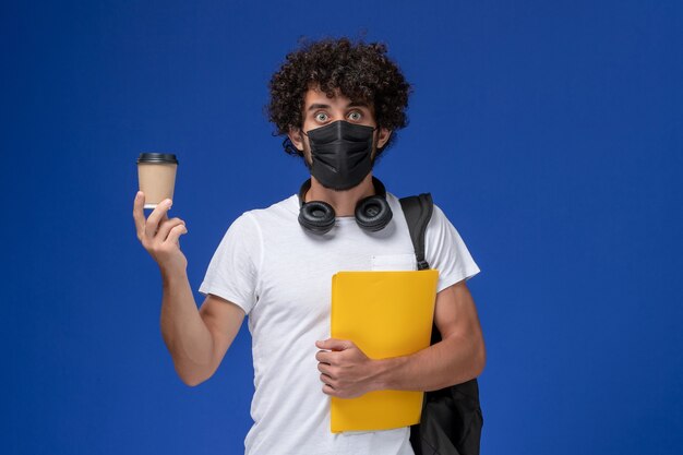 Giovane studente maschio di vista frontale in maglietta bianca che porta maschera nera e che tiene caffè file gialli su sfondo blu.