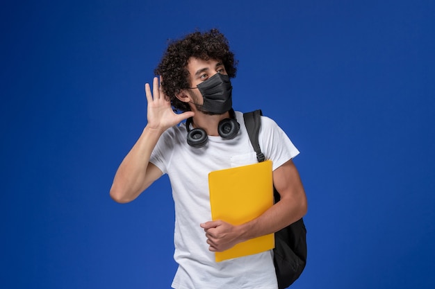 Giovane studente maschio di vista frontale in maglietta bianca che porta maschera nera e che tiene file gialli cercando di sentire sullo scrittorio blu chiaro.