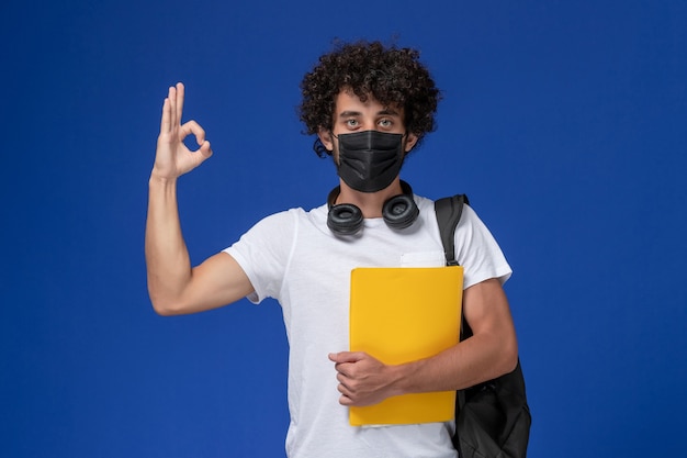 Giovane studente maschio di vista frontale in maglietta bianca che porta maschera nera e che tiene file gialli su sfondo azzurro.