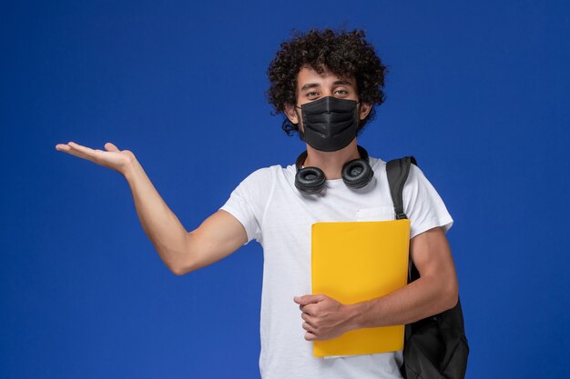 Giovane studente maschio di vista frontale in maglietta bianca che porta maschera nera e che tiene file gialli su sfondo azzurro.