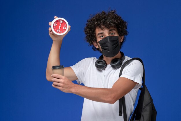 Giovane studente maschio di vista frontale in maglietta bianca che porta maschera nera e che tiene tazza di caffè con l'orologio sullo scrittorio blu.