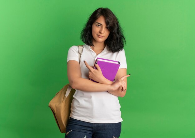 Giovane studentessa abbastanza caucasica fiduciosa che indossa i punti della borsa sul lato che tiene il libro sul verde con lo spazio della copia