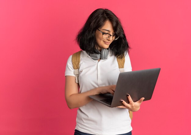 Giovane studentessa abbastanza caucasica soddisfatta con le cuffie sul collo con gli occhiali e la borsa posteriore tiene e guarda il laptop sul rosa con lo spazio della copia