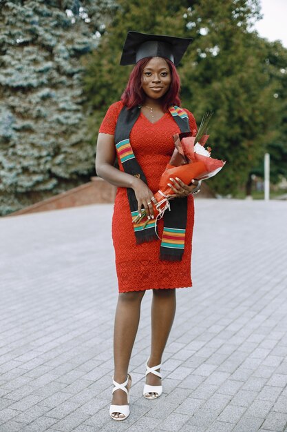 Giovane studentessa afroamericana vestita di abito rosso e berretto accademico. Ragazza in posa per una foto e con in mano un fiore