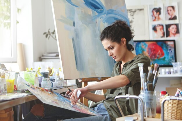 Giovane studentessa che ha lezioni allo studio d'arte, imparando a disegnare paesaggi, cercando di mescolare diversi acquerelli su cartone. Donna concentrata con i capelli scuri, vestita casualmente, pittura