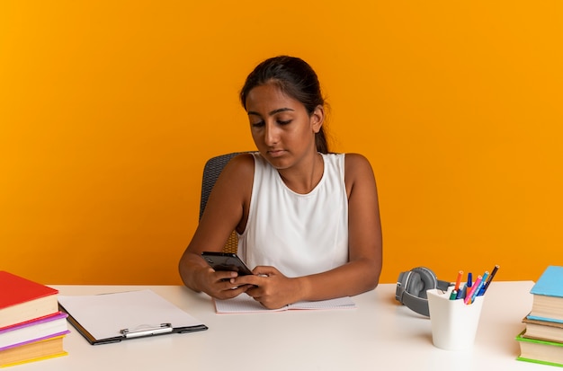 Giovane studentessa che si siede allo scrittorio con gli strumenti della scuola che tengono e che esaminano il telefono isolato sulla parete arancione