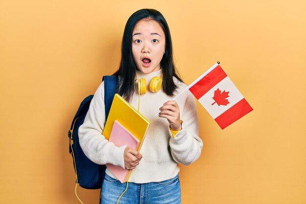 Giovane studentessa cinese di scambio di ragazze che tiene la bandiera del Canada impaurita e scioccata dalla sorpresa e dall'espressione stupita, dalla paura e dal viso eccitato.