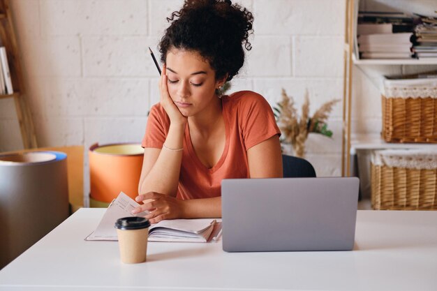 Giovane studentessa con i capelli ricci scuri seduta al tavolo con il laptop e una tazza di caffè per andare appoggiandosi a una mano studiando premurosamente a casa accogliente