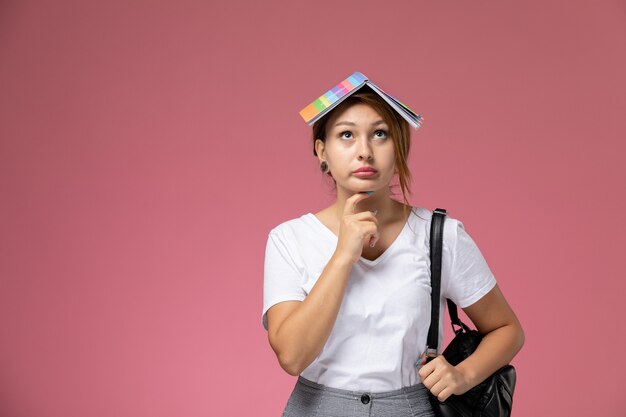 Giovane studentessa di vista frontale in maglietta bianca con il quaderno e la borsa che posa e che pensa sul libro di studio rosa di studio dell'università di lezione del fondo rosa